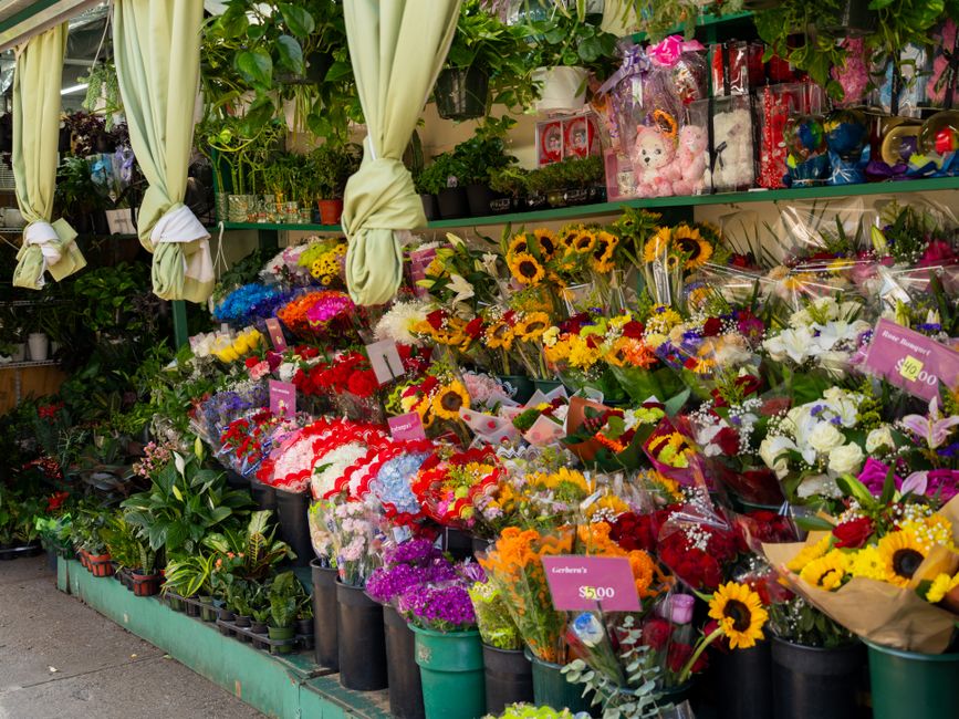 An outdoor flower stall displaying a vibrant selection of fresh floral arrangements, including bouquets of sunflowers, roses, lilies, and mixed seasonal flowers, wrapped in clear and colored cellophan
