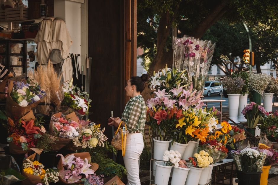 A woman with dark hair tied in a bun, dressed in a green and white checkered shirt and white pants, stands outside a florist shop during daytime, selecting various fresh flower bouquets and arrangemen