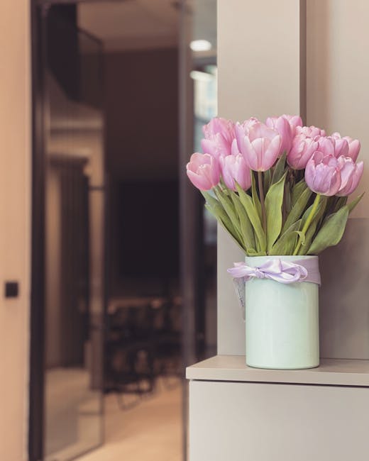 A bouquet of fresh pink tulips with green stems and leaves arranged in a white cylindrical ceramic vase tied with a light purple ribbon, placed on a white surface against a neutral wall. The tulips ar