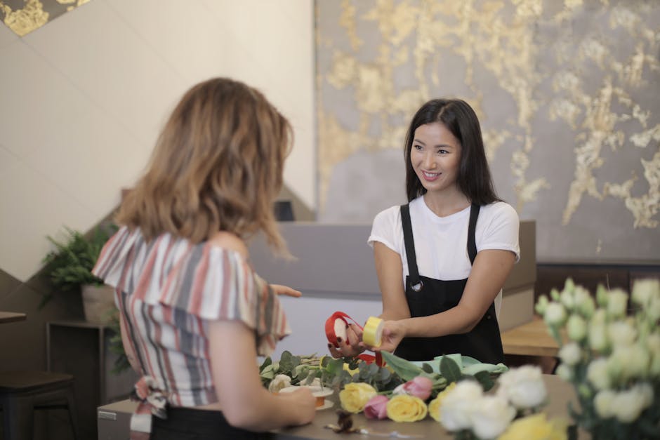 A florist at the Belmont flower shop engaged in a fresh flower arrangement for a floral gift, smiling as she hands over a small craft tape and floral ribbon to a customer. The arrangement features vib